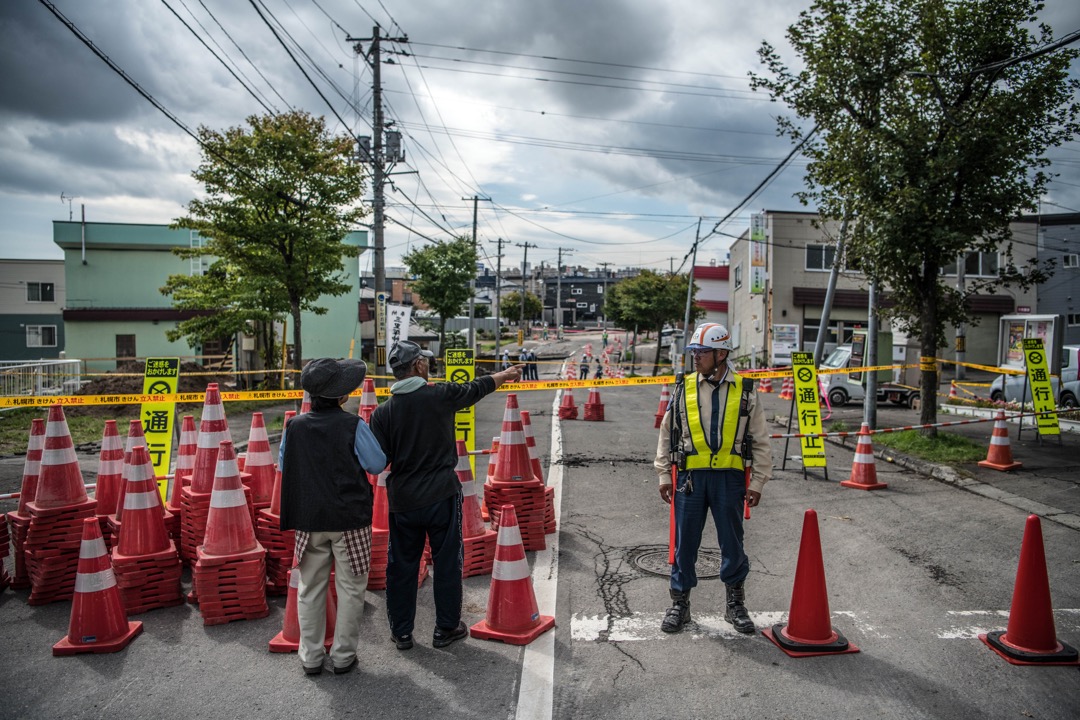 2018年9月8日,日本札幌清田区,道路在地震中严重损毁,需封闭进行维修,有道路工人在封锁线引导市民到安全地方,两名途人远观室内的损毁情况。