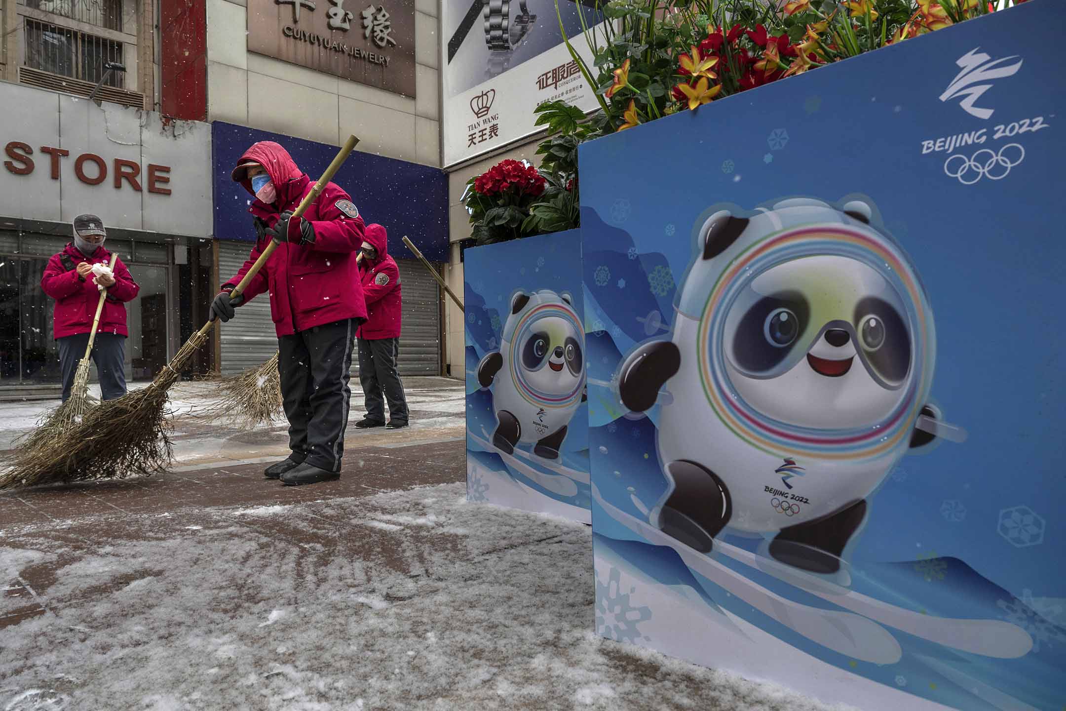 2022年1月20日北京,志愿者在北京冬奥会和残奥会海报旁边扫雪,海报展示了吉祥物“冰墩墩”。